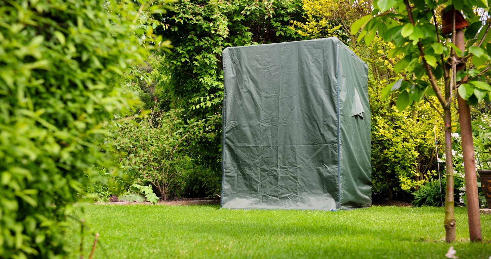 Ein mit einer grünen Plane bedecktes Objekt in einem Garten umgeben von Büschen und Bäumen.