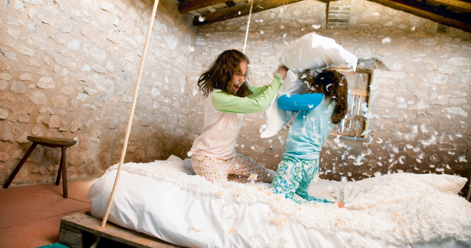 Zwei Kinder veranstalten eine Kissenschlacht auf einem Bett in einem rustikalen Zimmer, um sie herum fliegen Federn.